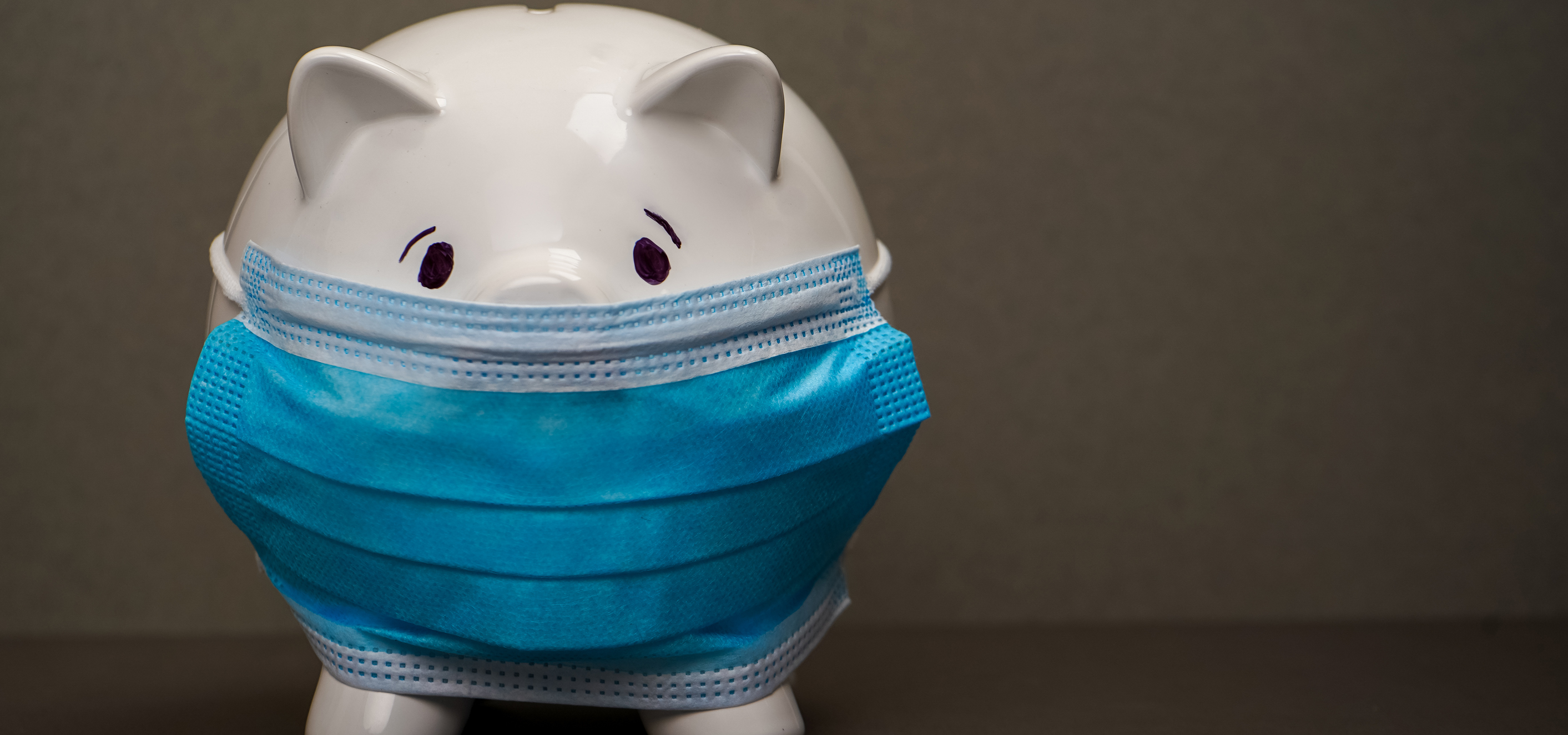A quintessential white piggy bank left of center wearing a blue, coronavirus type mask, in front of a brownish background.